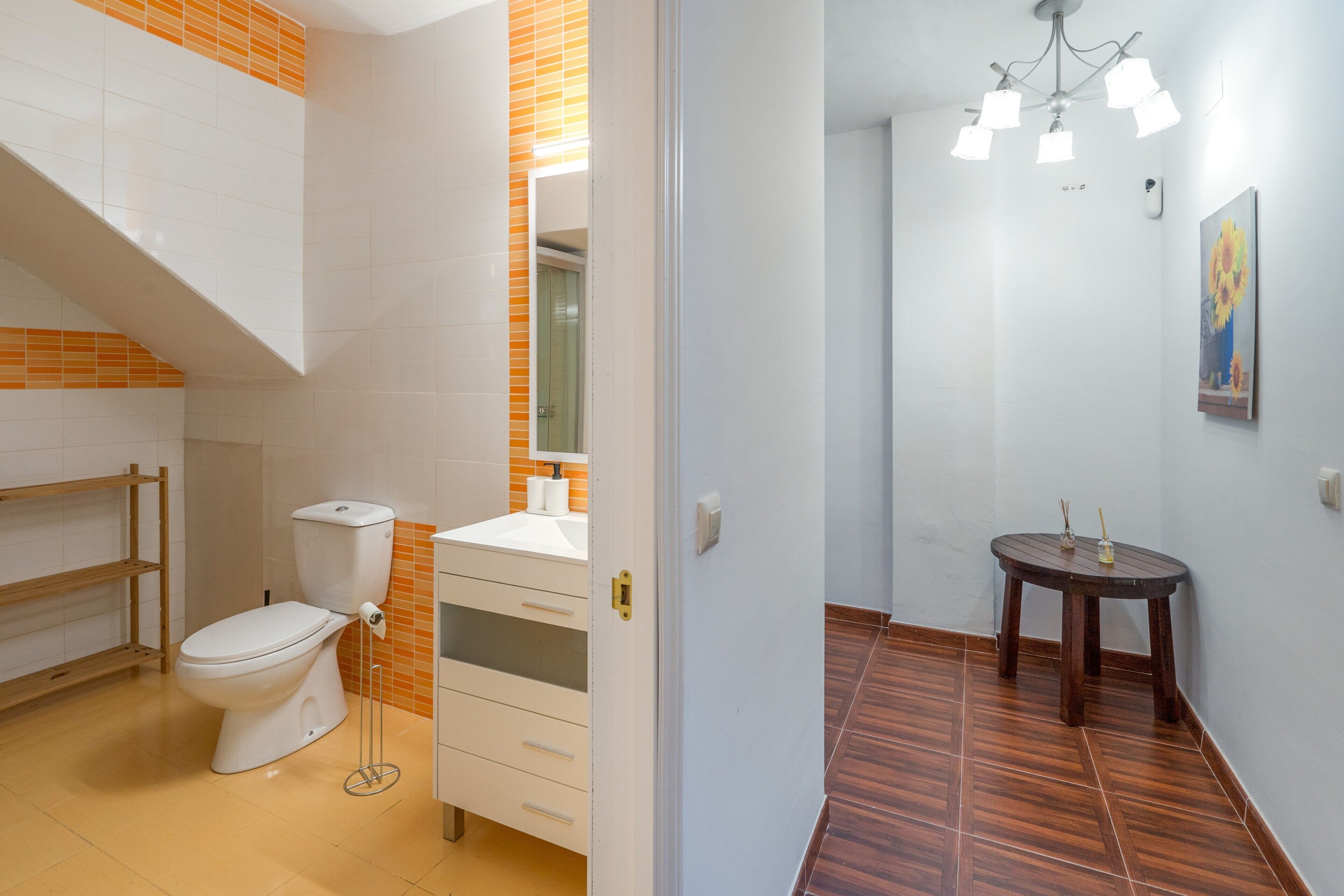 Baño con inodoro, lavabo y espejo. Se observa una repisa de madera y una pequeña mesa con un florero.