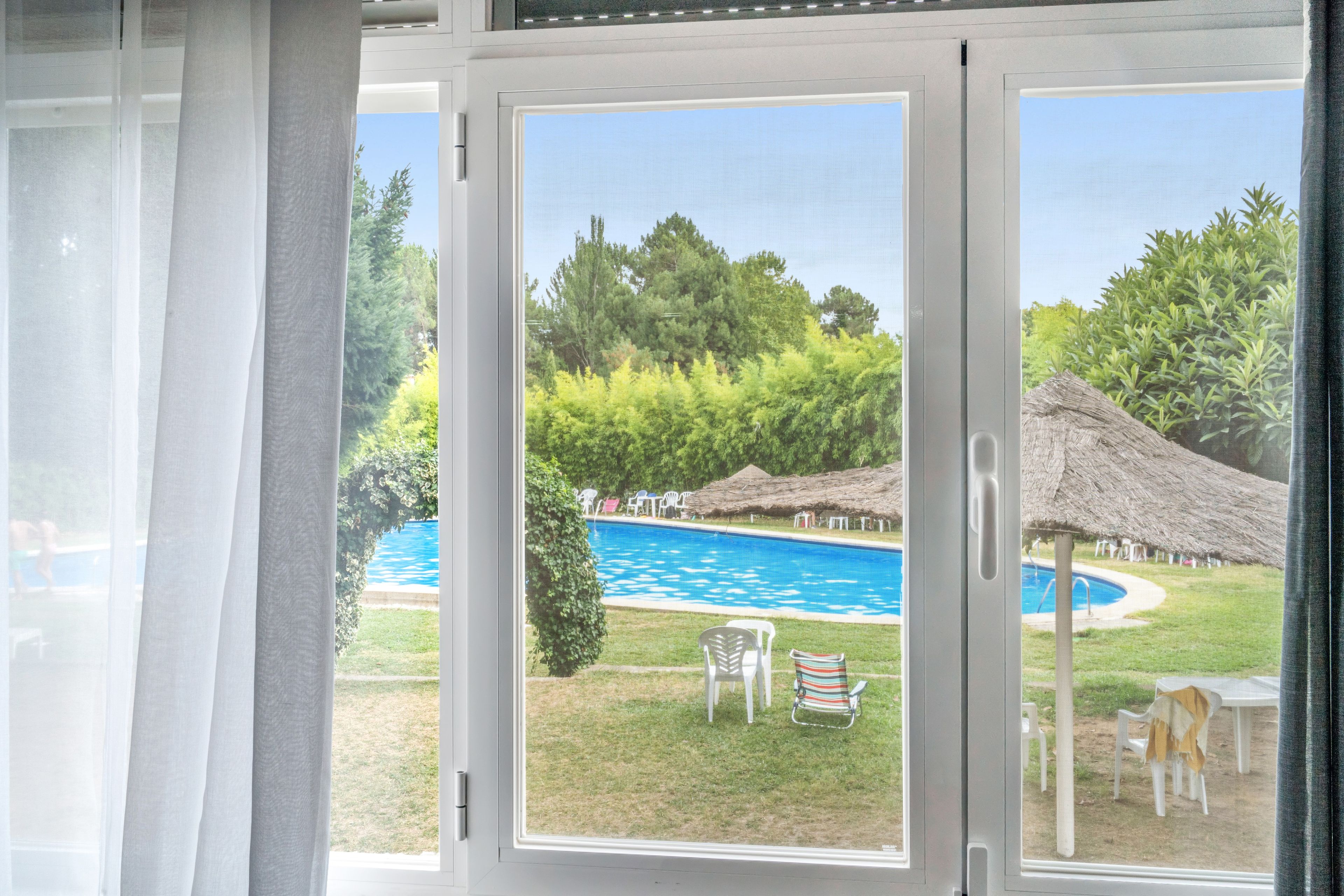 Une vue à travers une fenêtre donnant sur une piscine, des chaises et une structure à toit de chaume.