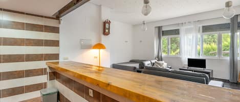 A modern kitchen with a wooden countertop, a built-in trash bin, and a dining area with a sofa and a television.