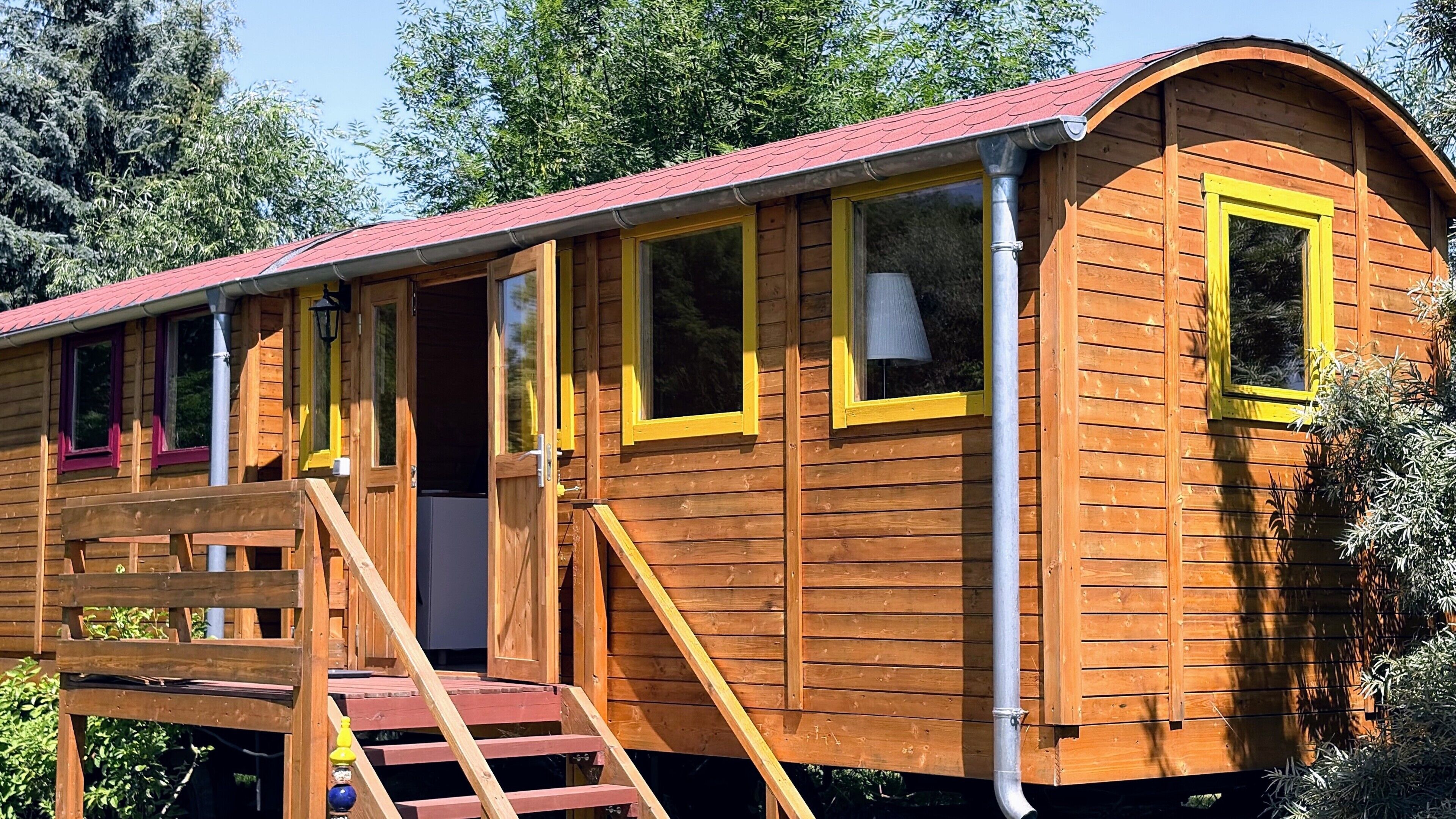 Eine Holzhütte mit rotem Dach, mehreren Fenstern mit gelben Rahmen und einer Holztreppe, die zum Eingang führt.