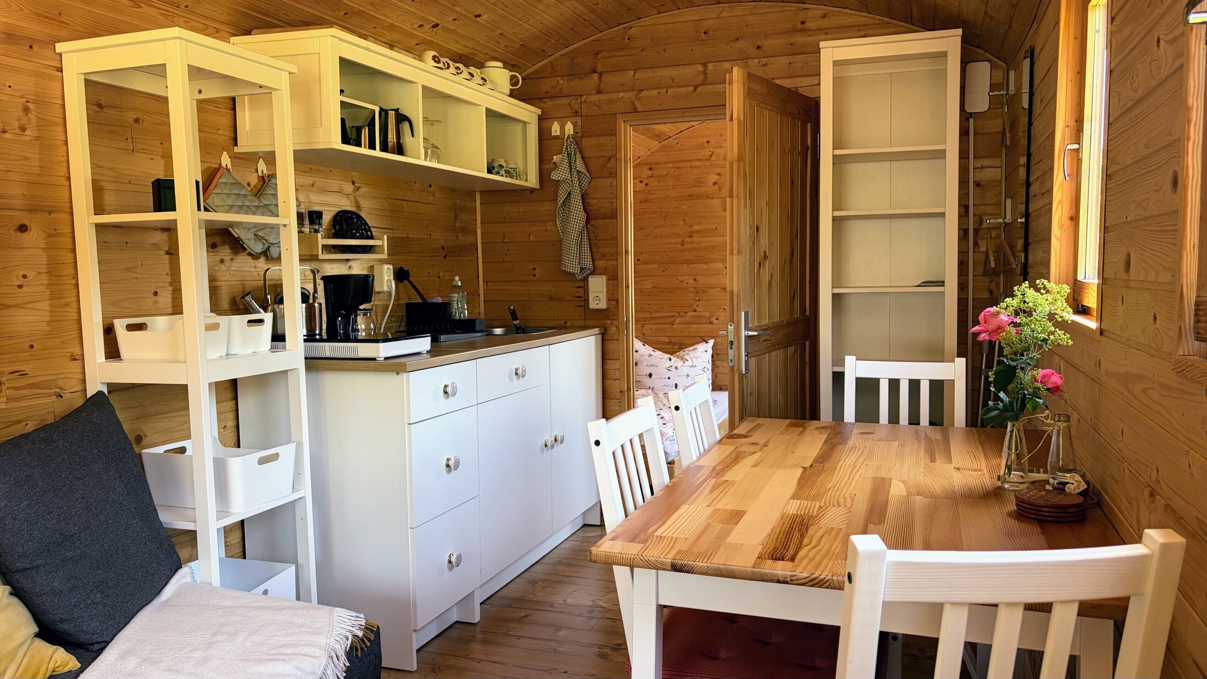 A wooden interior with a kitchen area, white cabinets, a dining table with chairs, and a vase with flowers.