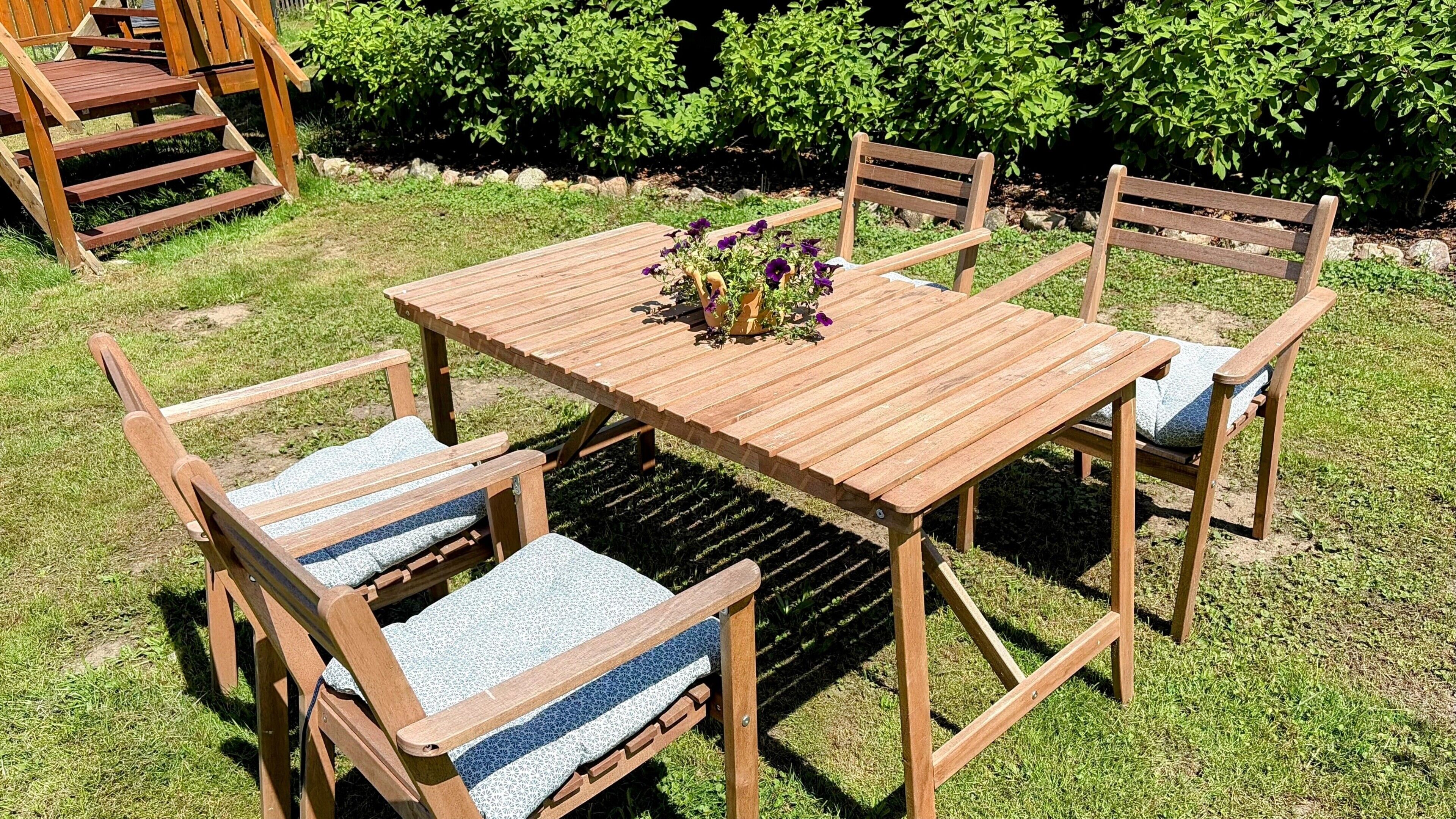 A wooden outdoor table with chairs, surrounded by a grassy area and bushes.