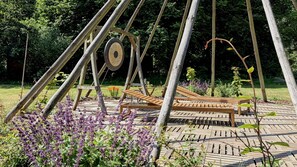 A wooden swing set with a bench, surrounded by a garden with purple flowers and a paved area.