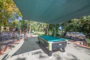 Et udendørs poolbord under et overdækket område med tilhørende borde og stole, beliggende i en parklignende omgivelser med træer og et fjerntliggende udsyn til en golfbane.