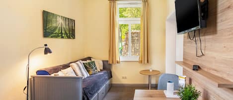 A compact living space with a sofa, a small table, a television mounted on the wall, and a window with curtains.