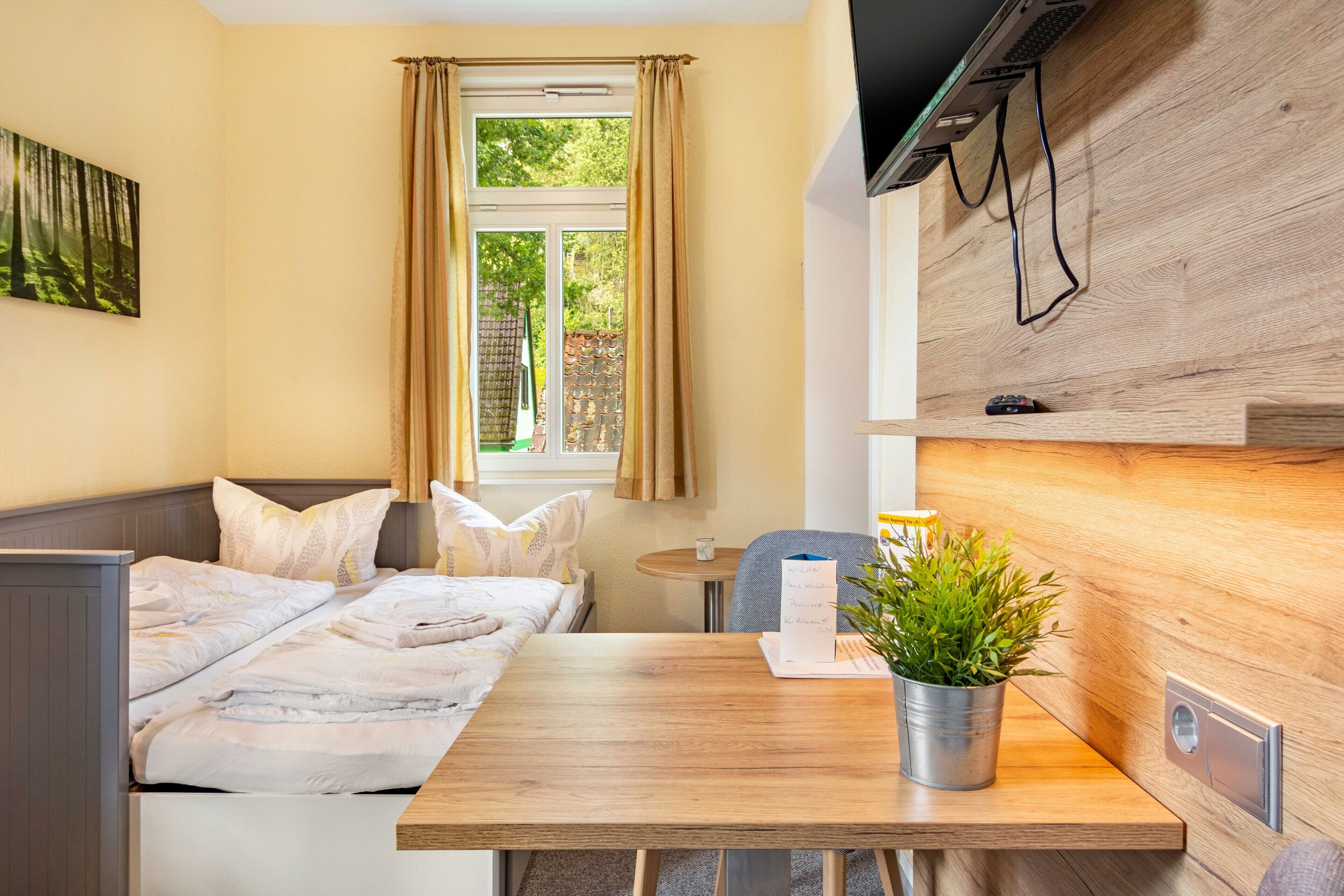Ett hotellrum med en säng, ett litet bord med en växt, ett fönster med gardiner och en tv som är monterad på väggen.