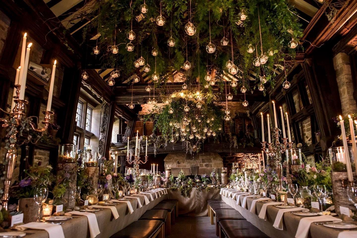 Una larga mesa de comedor con centros de mesa iluminados por velas, rodeada de altos candelabros, en una habitación con vigas de madera a la vista y orbes de vidrio colgantes.