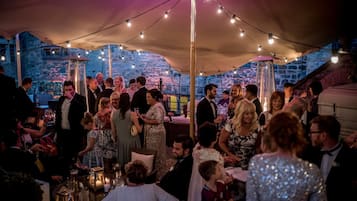 A tented outdoor event with people mingling, tables with drinks, and a stone building in the background.