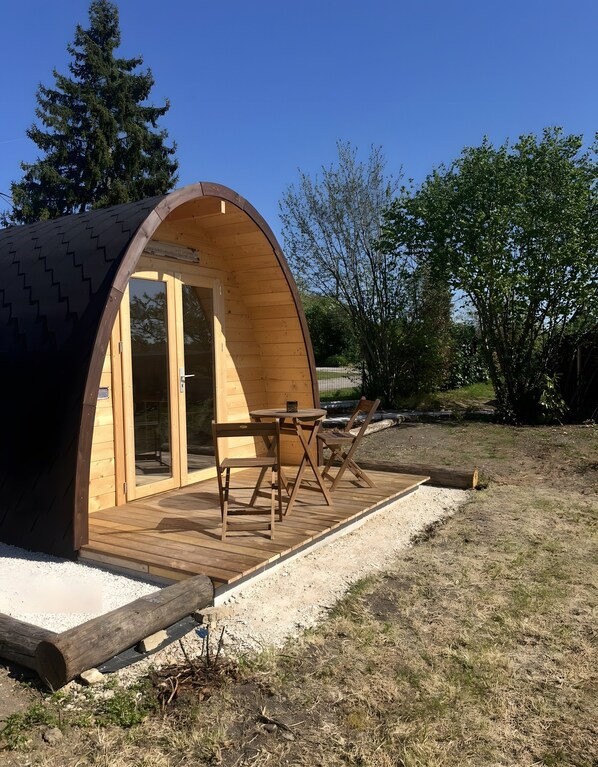 A wooden dome-shaped structure with a table and chairs outside, surrounded by trees and gravel.