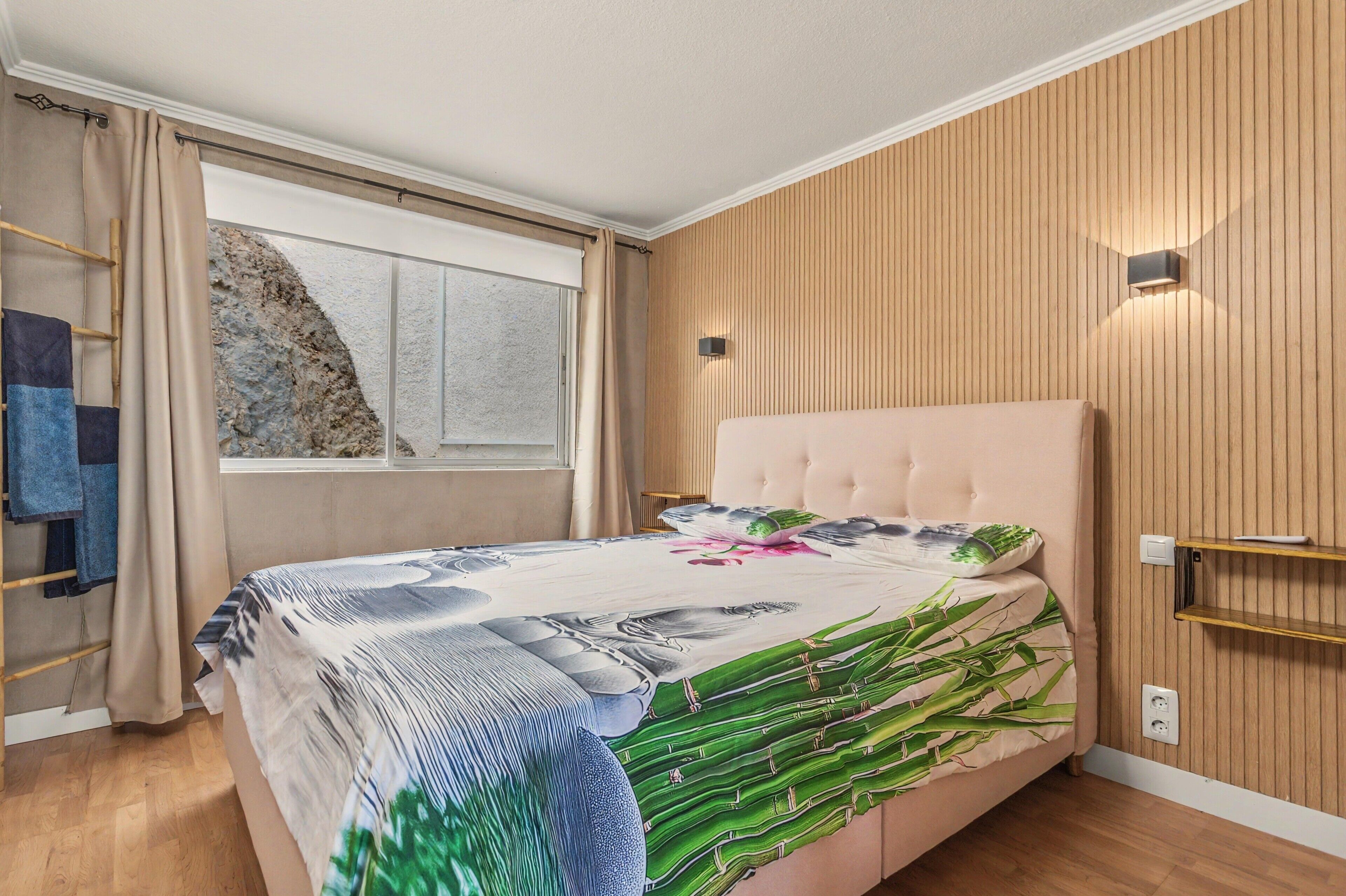 A bedroom with a bed, a window, wooden panel walls, and a wall-mounted light fixture.