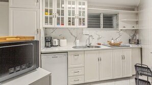 Une cuisine compacte avec des meubles blancs, un lave-vaisselle encastré, une machine à café et une fenêtre avec des stores.