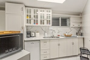 A compact kitchen with white cabinets, a built-in dishwasher, a coffee maker, and a window with blinds.