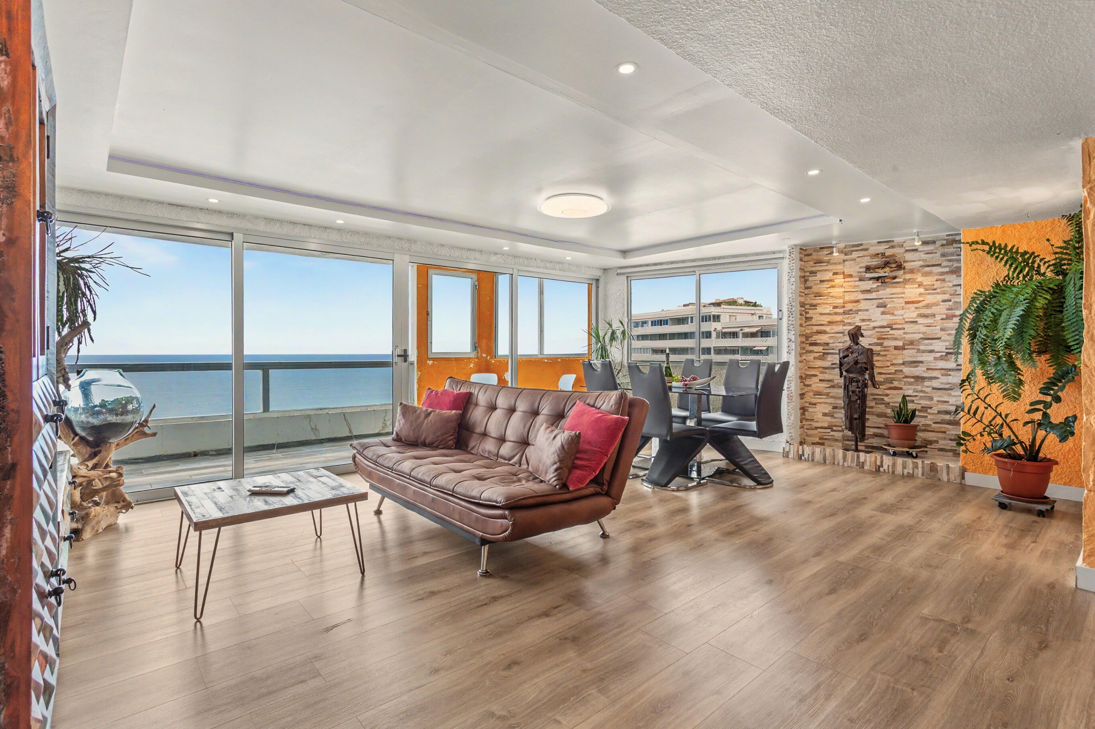 Amplia sala de estar con un ventanal que da al mar, un sofá de cuero, una mesa de comedor con sillas y una pared decorativa con un reloj.