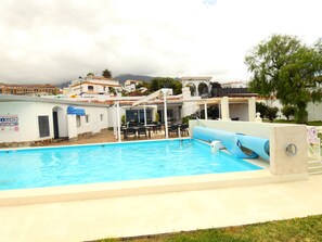 A swimming pool with a sun lounger and a covered seating area, surrounded by buildings and a cloudy sky. - Vacation Apartment 'Ciel et Mer' with Sea View, Wi-Fi, and Air Conditioning (Los Realejos)