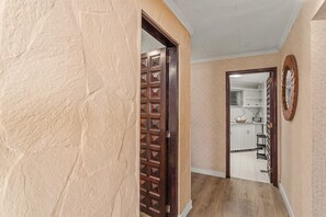 A hallway with a wooden door, a kitchen area with cabinets, and a tiled floor.