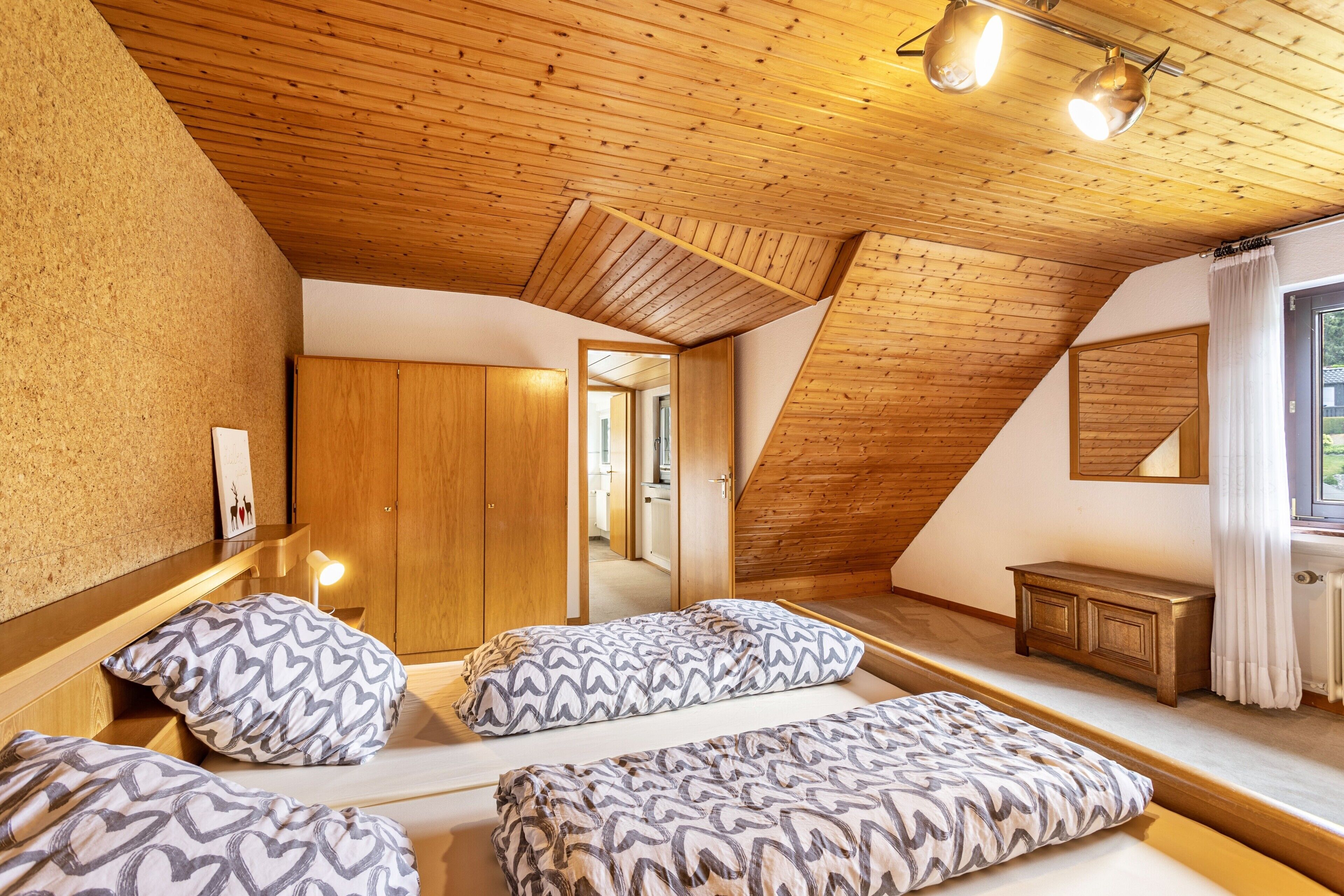 Un dormitorio con techo inclinado, paredes de madera, cama con ropa de cama estampada, mesita de noche y espejo.