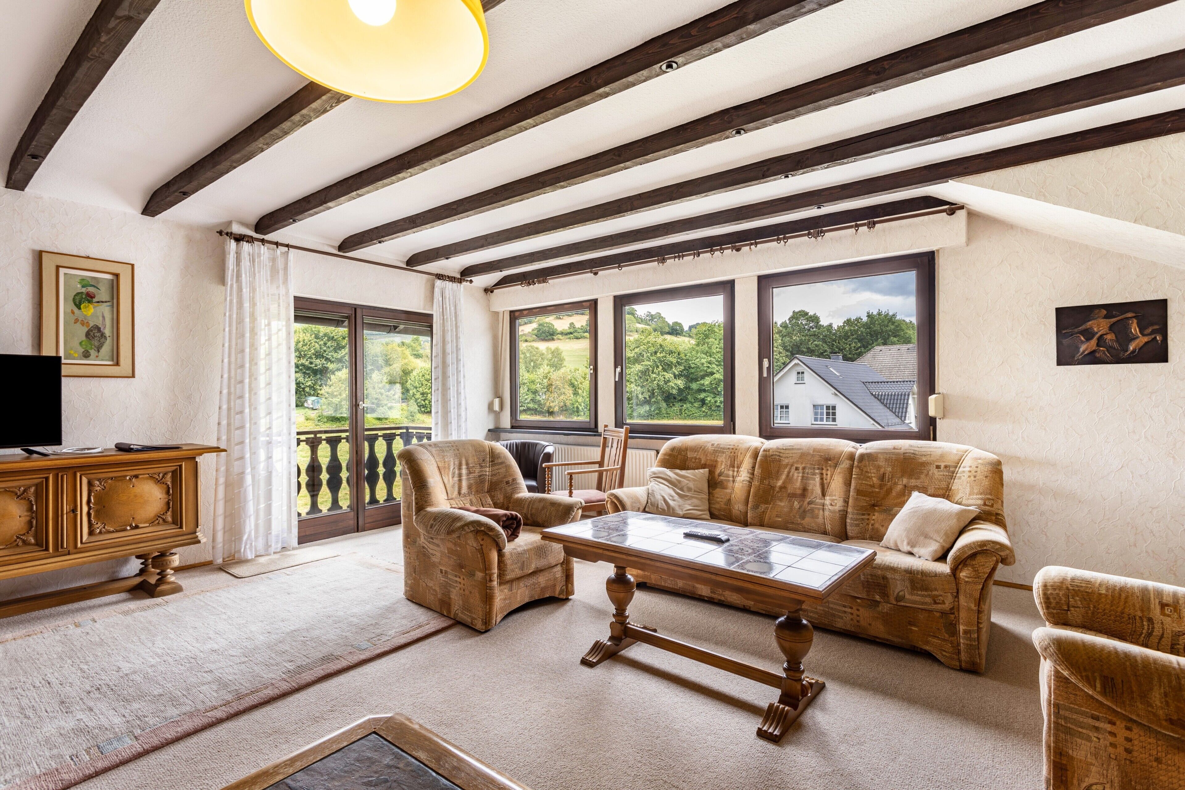 A spacious living room with a high ceiling, exposed wooden beams, a large window with a view of greenery, a sofa, a coffee table, and a television on a wooden cabinet.
