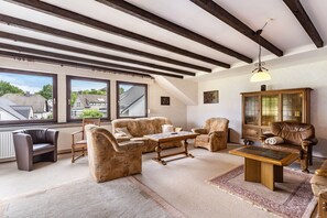 A spacious living room with a high ceiling, large windows, and a view of the outside. There is a fireplace, a wooden cabinet, and a variety of seating including a sofa, armchairs, and a table. - Apartment 'Fewo Wrede Am Sauerland-Radring 2' with Mountain View, Balcony and Wi-Fi (Eslohe)