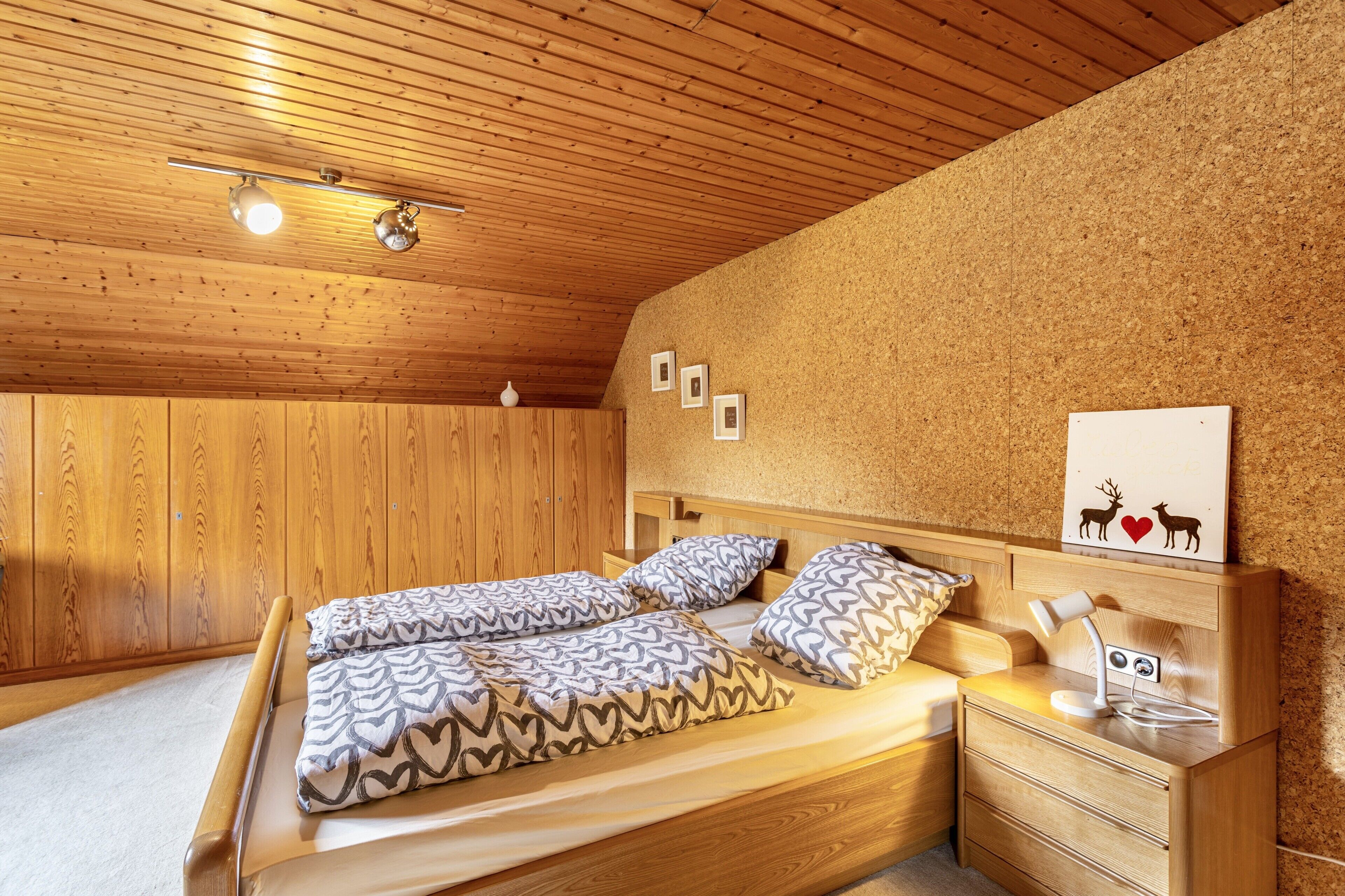 A bedroom with a wooden ceiling, a bed with patterned bedding, a bedside table with a lamp, and a corkboard wall with framed pictures.