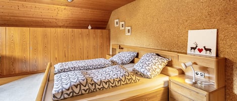 A bedroom with a wooden ceiling, a bed with patterned bedding, a bedside table with a lamp, and a corkboard wall with framed pictures.