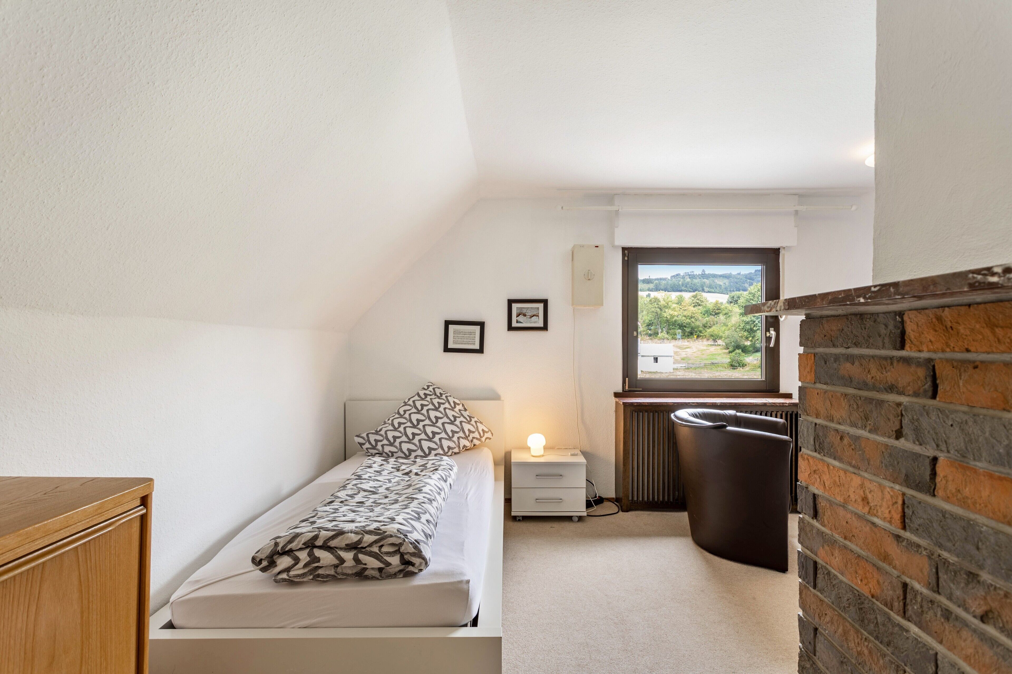 A small, well-lit bedroom with a single bed, a nightstand, a radiator, and a window with a view of greenery.