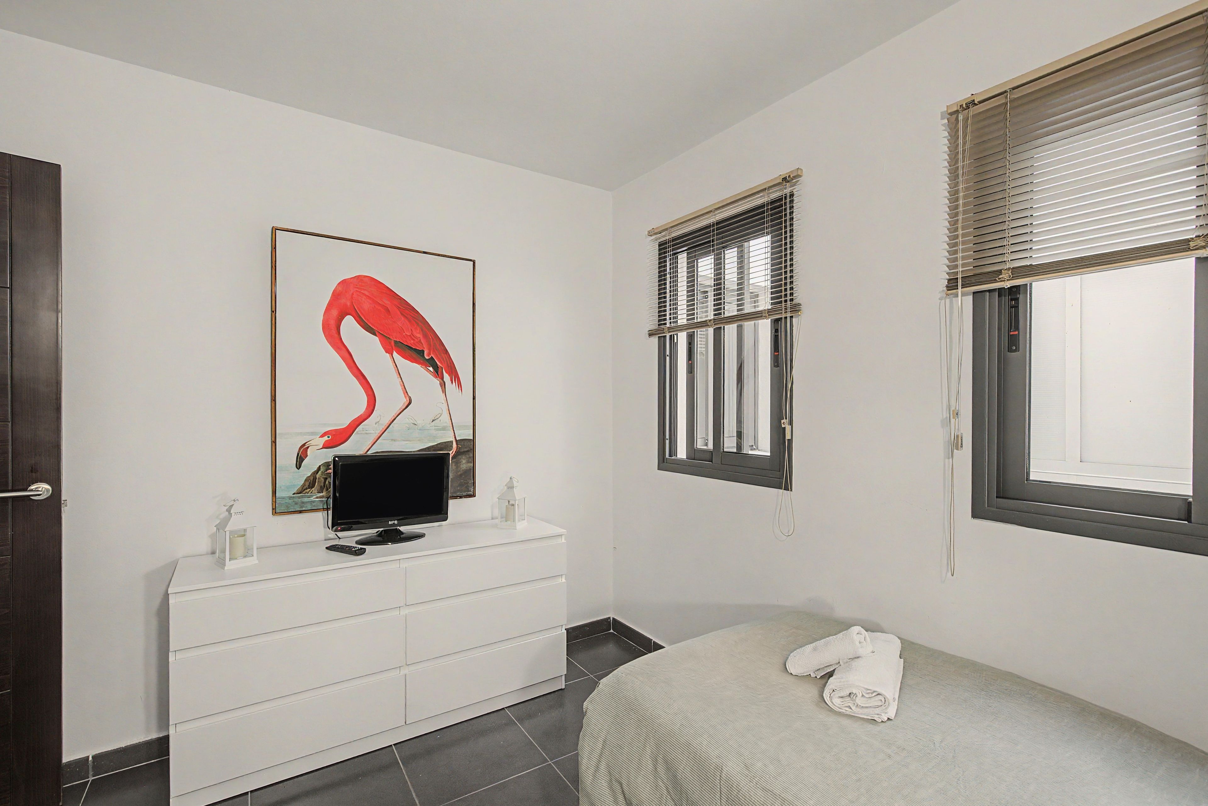 Ein Schlafzimmer mit einem Bett, einem Fernseher auf einer Kommode, einem Fenster mit Jalousien und einem Gemälde an der Wand.