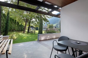 Une terrasse couverte avec une table et des chaises, un banc en bois et une vue sur un jardin luxuriant et des montagnes.