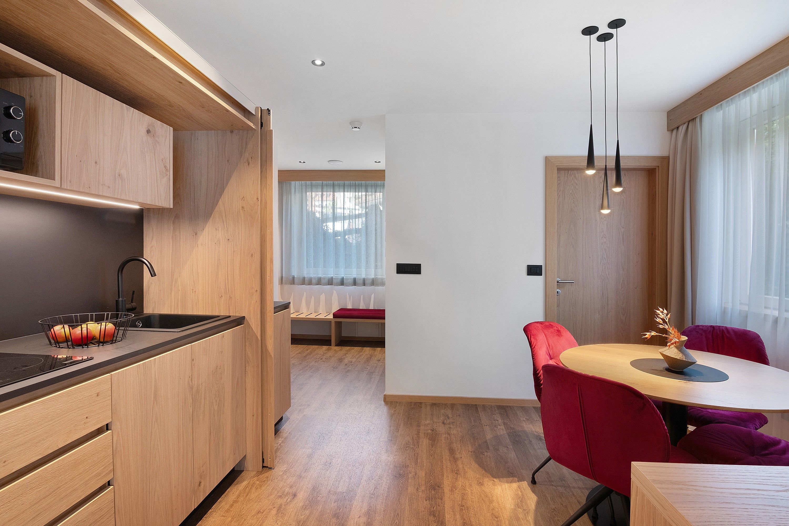Una cocina moderna con horno empotrado, un lavamanos y una cesta de frutas. Hay una zona de comedor con una mesa redonda y sillas rojas.