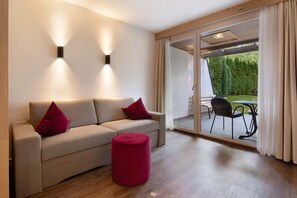Een moderne woonkamer met een bank, rode kussens, een rode poef en een glazen deur die leidt naar een terras met een tafel en stoelen.
