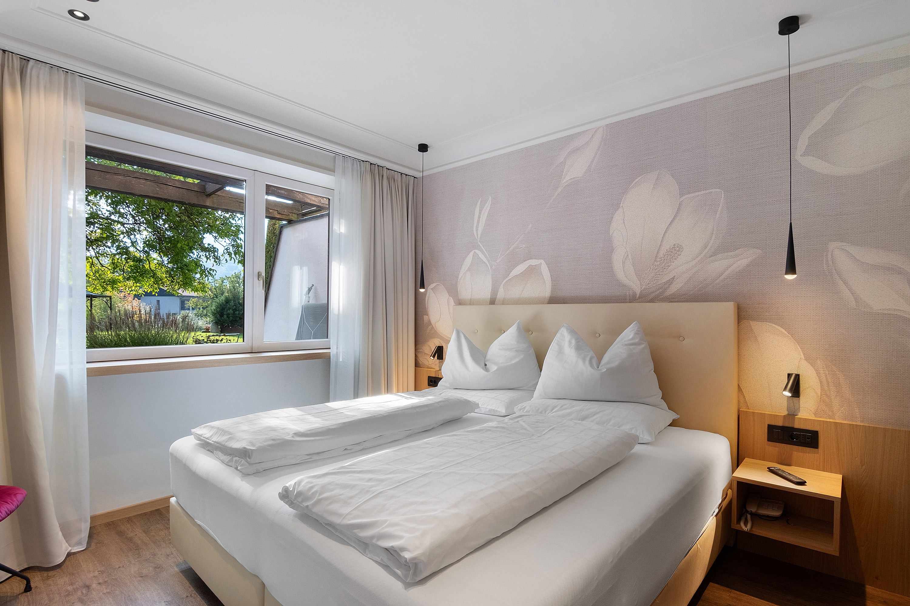 A hotel room with a large bed, a window with curtains, a wall with a floral pattern, and a bedside table with a lamp.