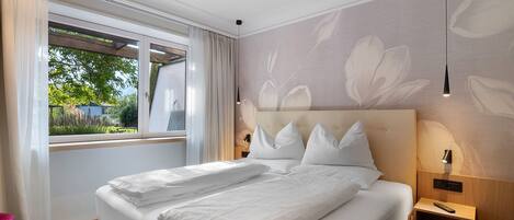 A hotel room with a large bed, a window with curtains, a wall with a floral pattern, and a bedside table with a lamp.