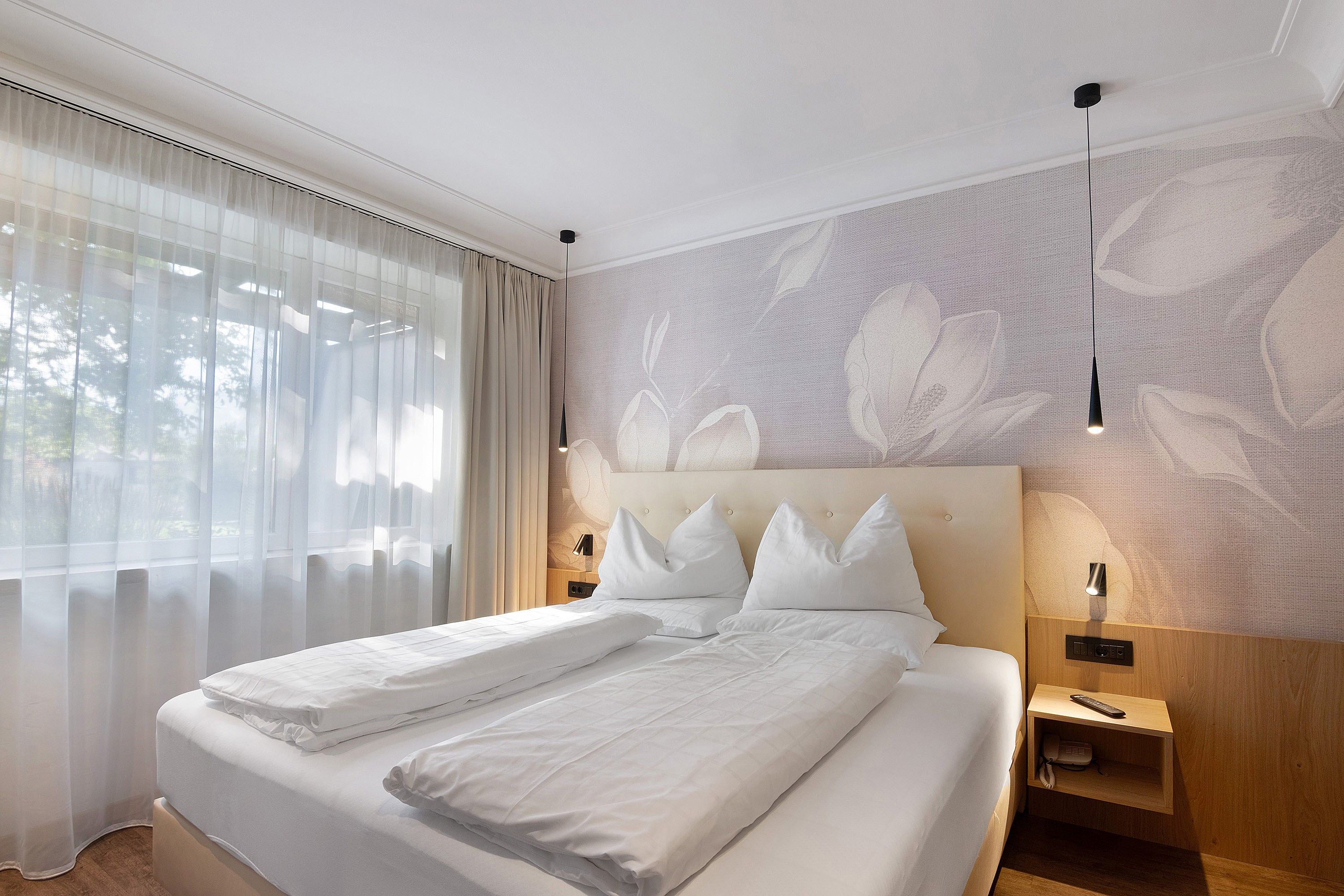 Ein Hotelzimmer mit einem großen Bett, zwei Nachttischen, einer Wandlampe und durchscheinenden Vorhängen.