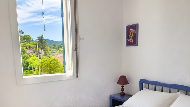 Une chambre à coucher avec un lit, une table de chevet, une lampe, une fenêtre donnant sur un paysage verdoyant et un tableau encadré au mur.