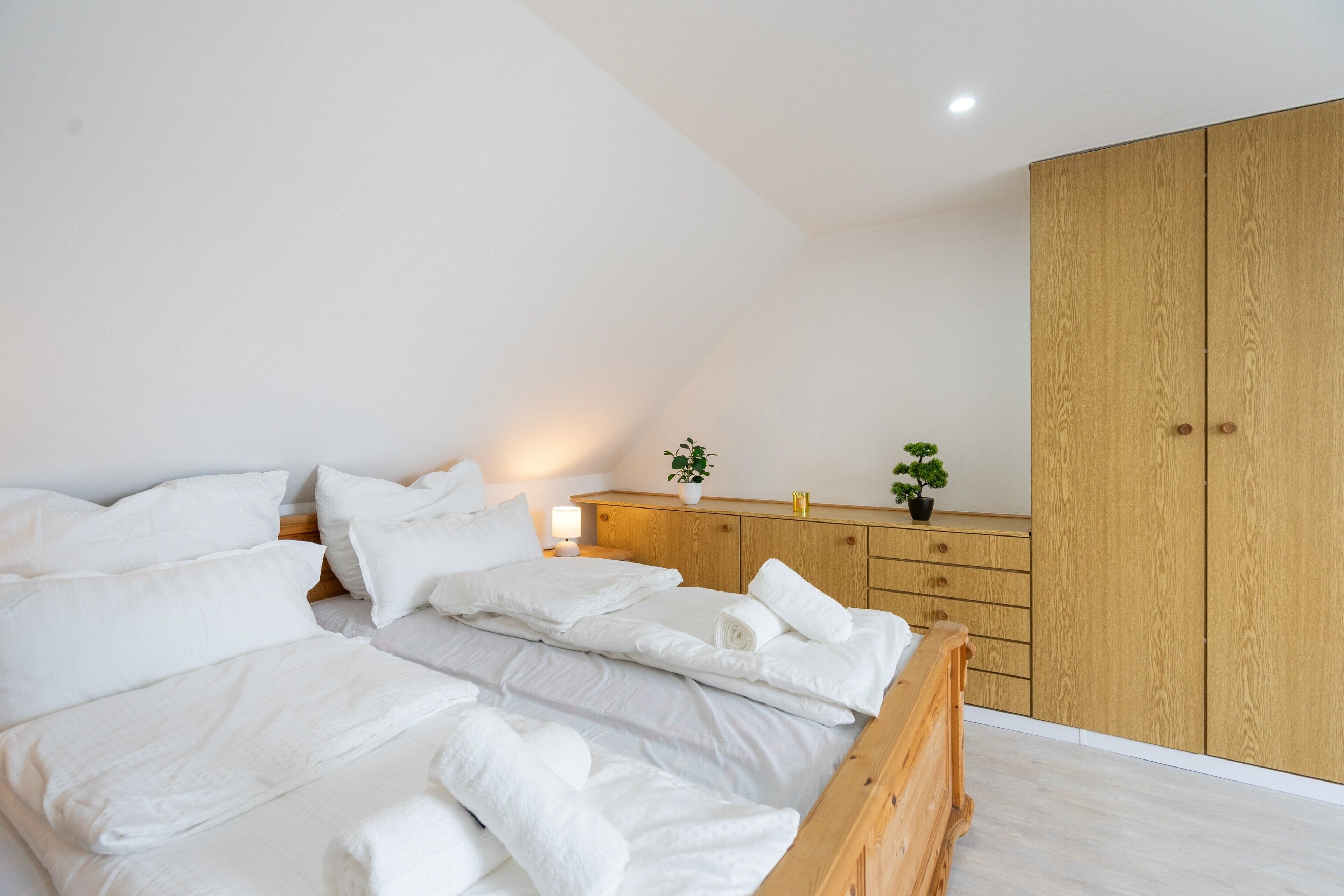 A bedroom with a bed, pillows, a nightstand, a lamp, a small plant, and a wooden wardrobe.