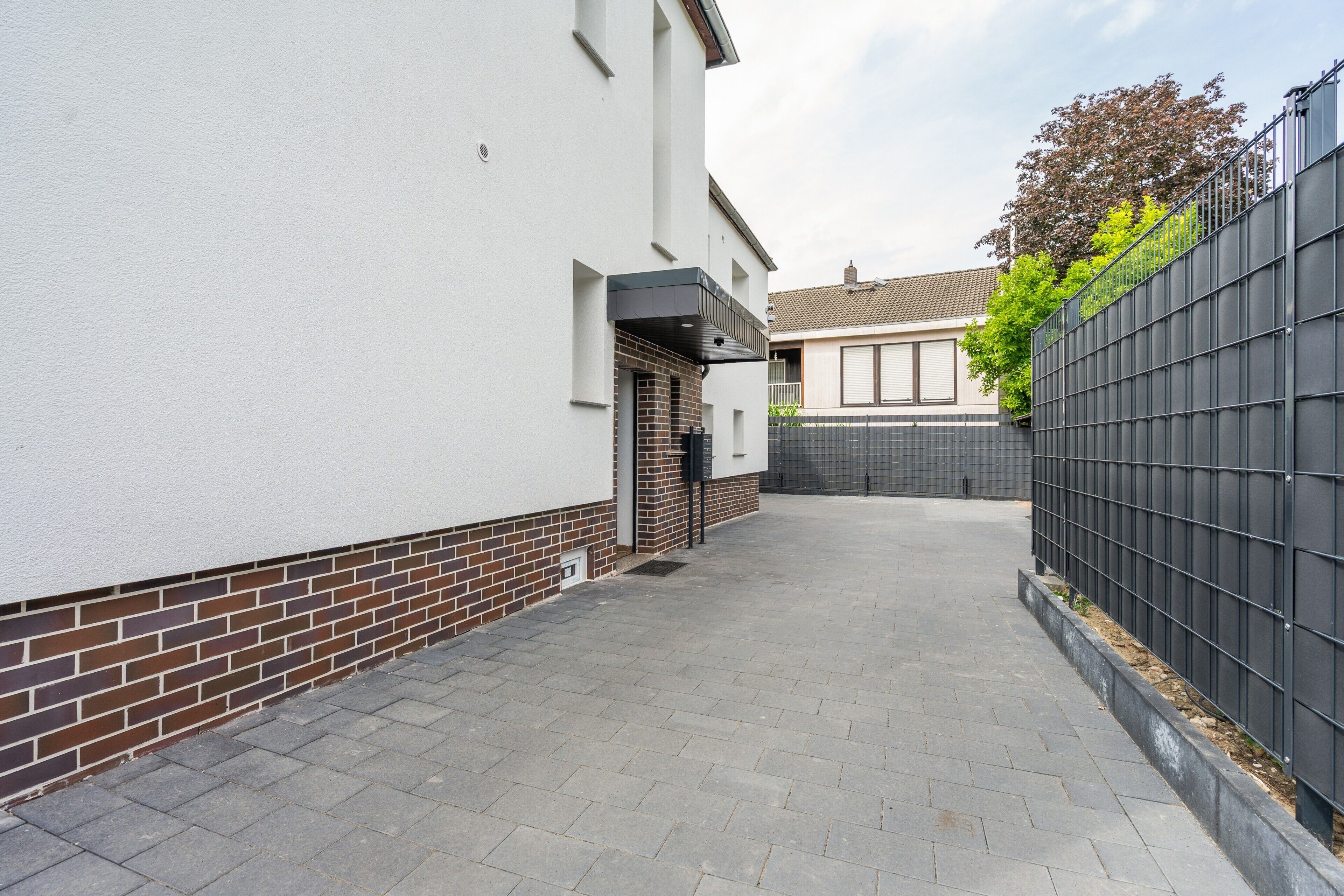 A narrow paved pathway leading to a residential entrance with a white wall, a brick base, and a black metal fence on one side.