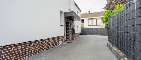 A narrow paved pathway leading to a residential entrance with a white wall, a brick base, and a black metal fence on one side.