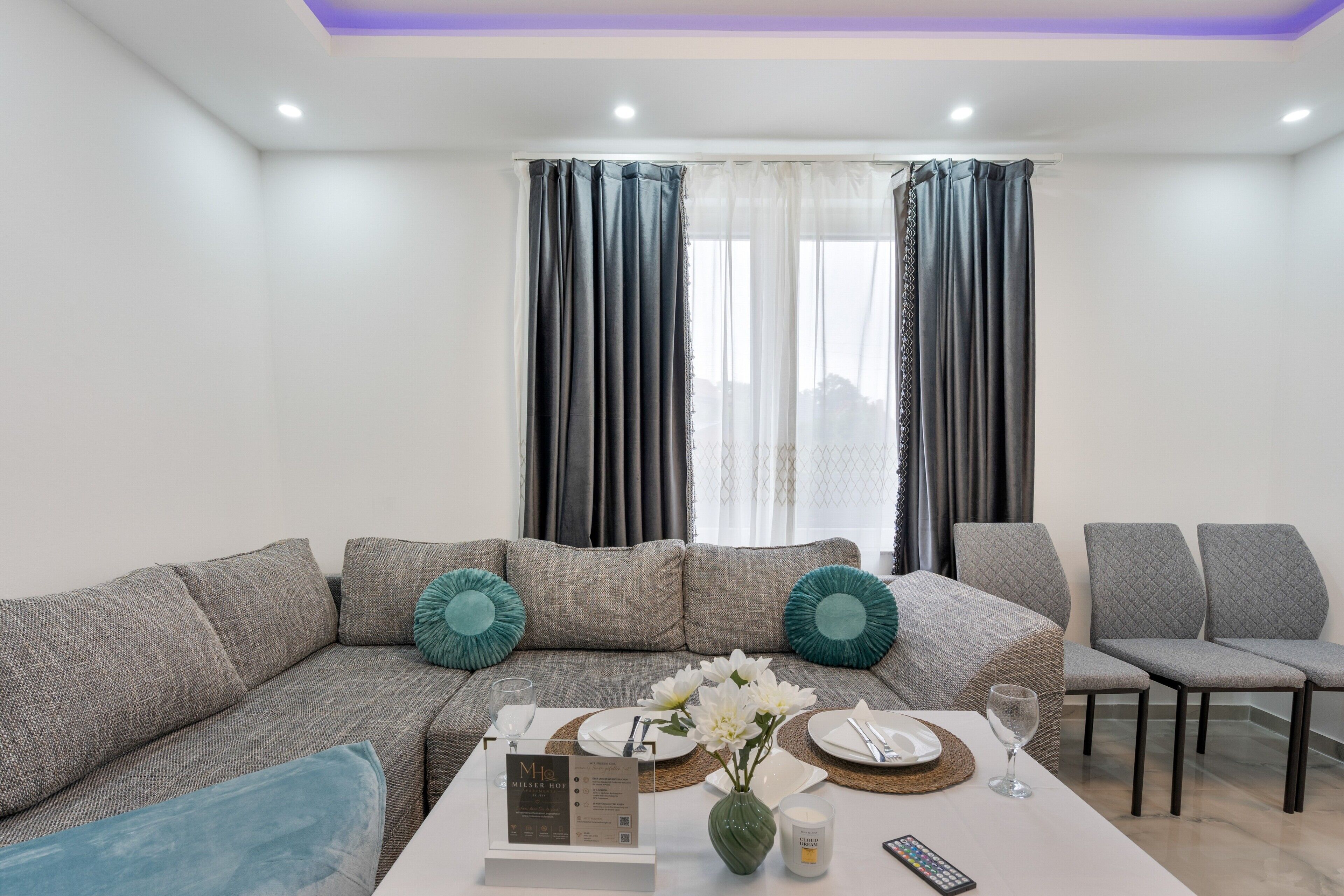 A modern living room with a sectional sofa, a coffee table, and a dining area set with plates and glasses.