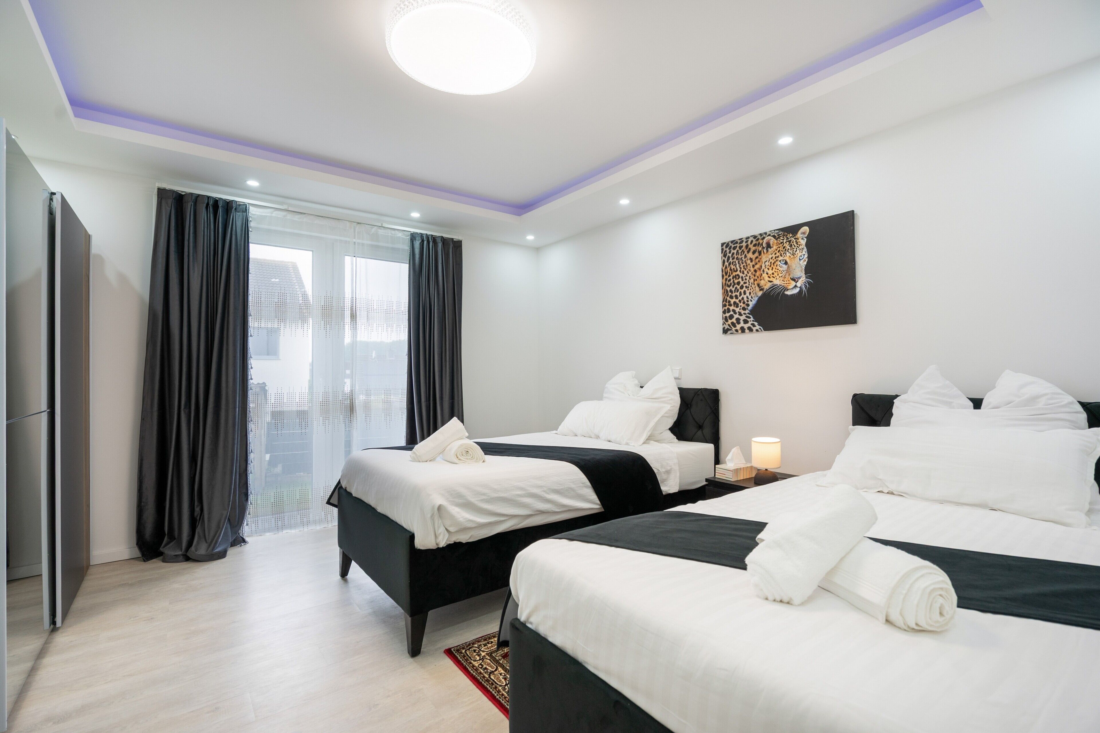 A hotel room with two beds, a large window with curtains, a painting on the wall, and a ceiling light.