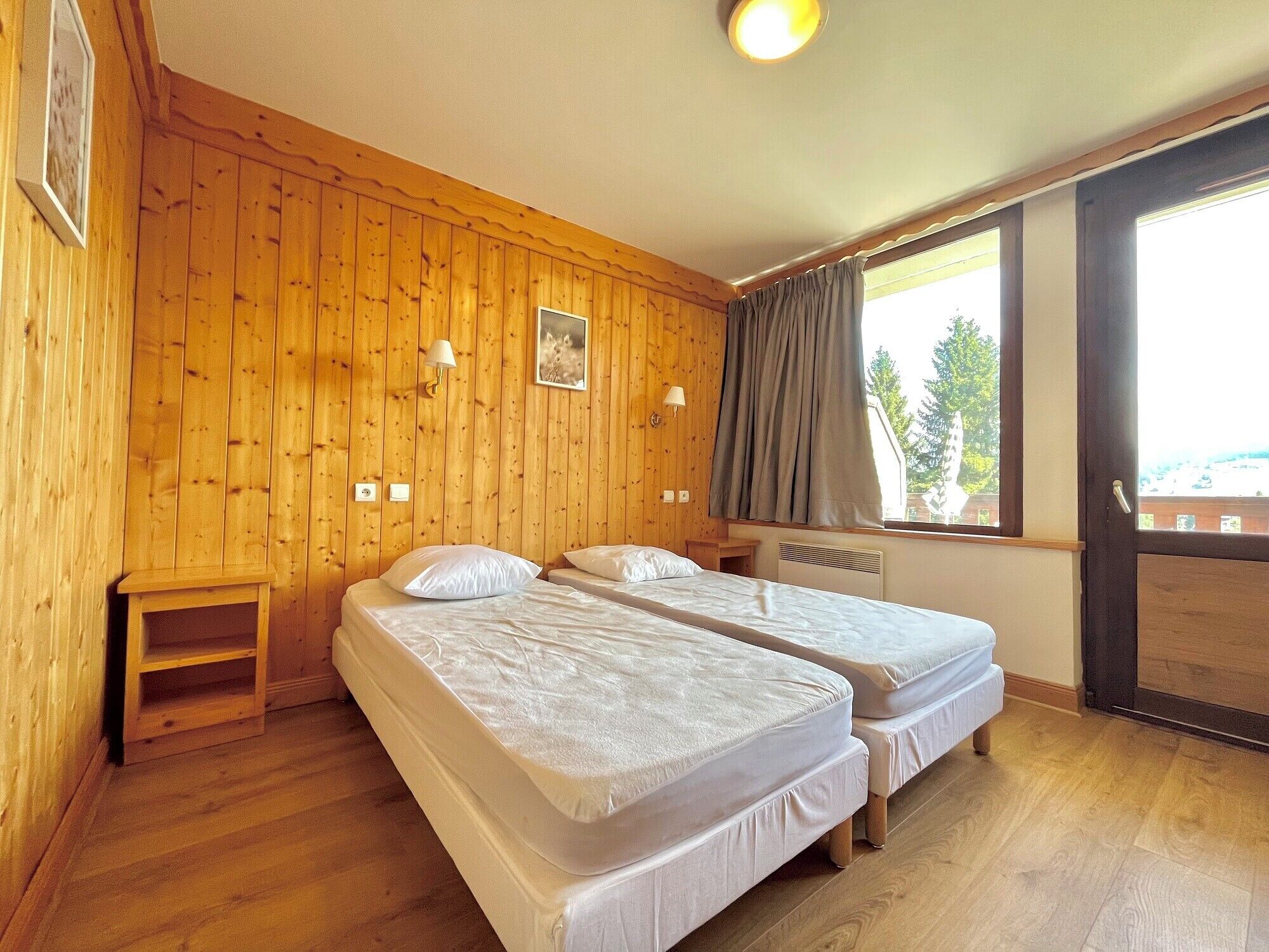 A room with wooden panel walls, two beds with white bedding, a small wooden nightstand, a window with curtains, and a ceiling light.