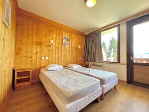 Una stanza con pareti in legno, due letti singoli con lenzuola bianche, un piccolo comodino in legno, una finestra con vista sul verde e una luce al soffitto.