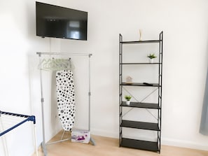 Habitació amb televisió penjada a la paret, un tendal amb taula de planxar i un moble prestatge metàllic negre amb objectes decoratius.