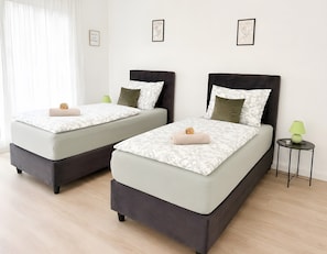 Two twin beds with dark headboards and light bedding, a small bedside table with a lamp, and a wall with framed artwork.