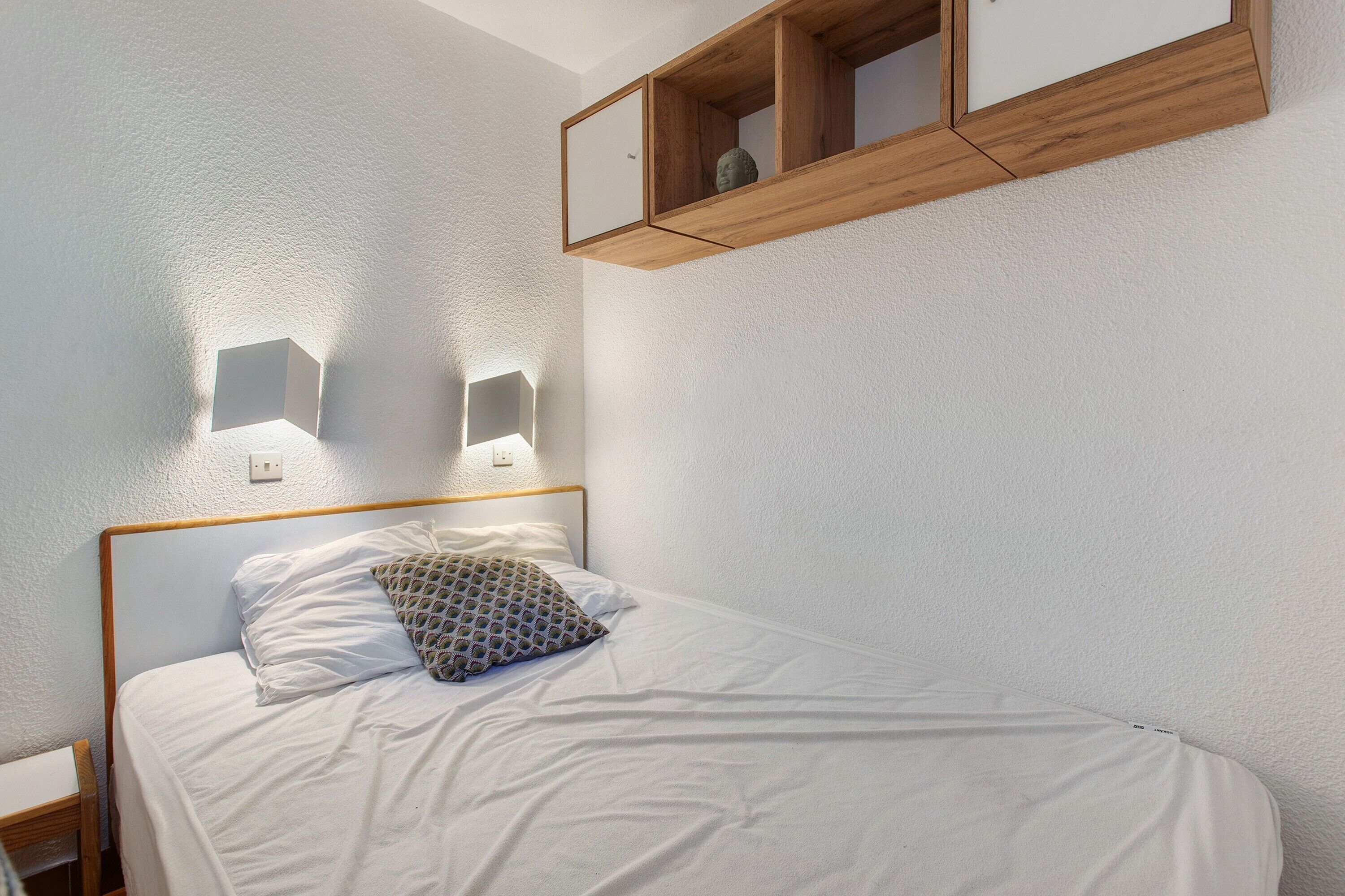 Un lit bien fait, agrémenté d’un coussin à motifs, d’une tête de lit en bois et d’une étagère murale sur laquelle repose un petit objet.