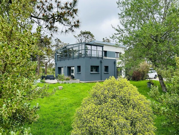 A modern two-story house with a balcony, surrounded by lush greenery and a well-maintained lawn.