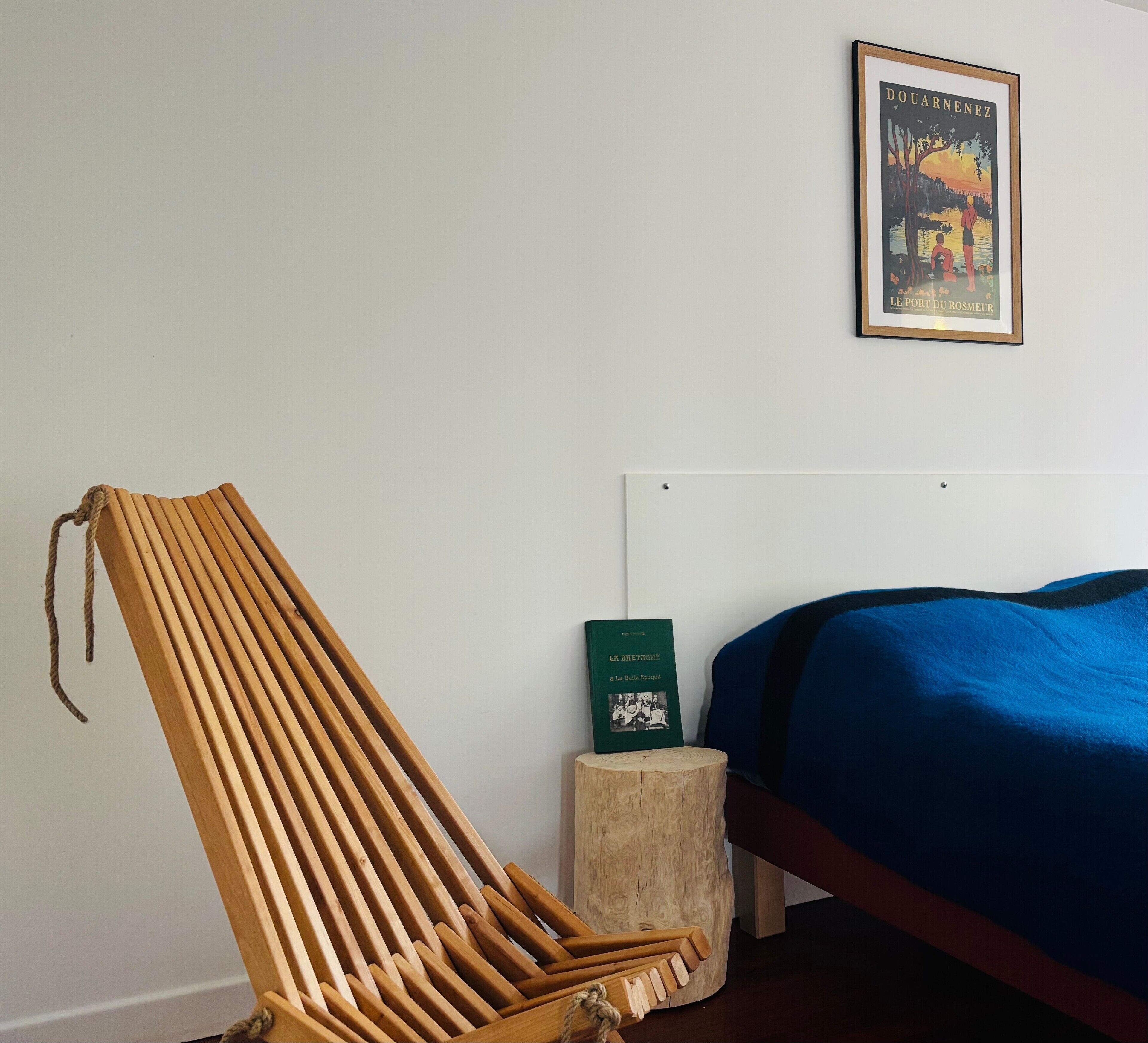 Een kamer met een houten stoel, een bed met een blauwe deken, een klein houten krukje en een lijst met een afbeelding aan de muur.