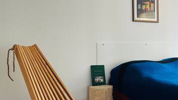 Een kamer met een houten stoel, een bed met een blauwe deken, een klein houten krukje en een lijst met een afbeelding aan de muur.