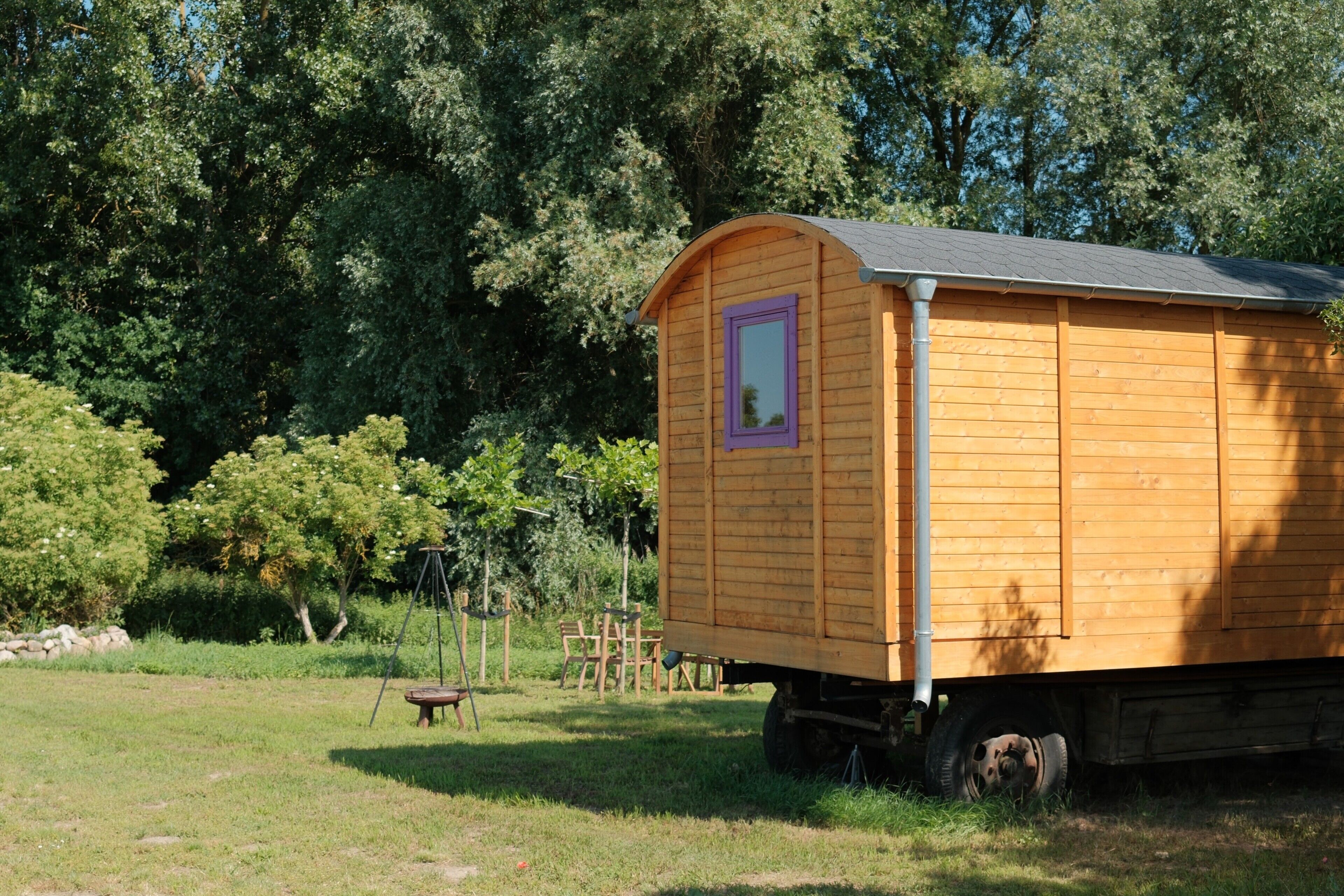 A wooden mobile home with a curved roof and a small window, situated in a grassy area with trees in the background.