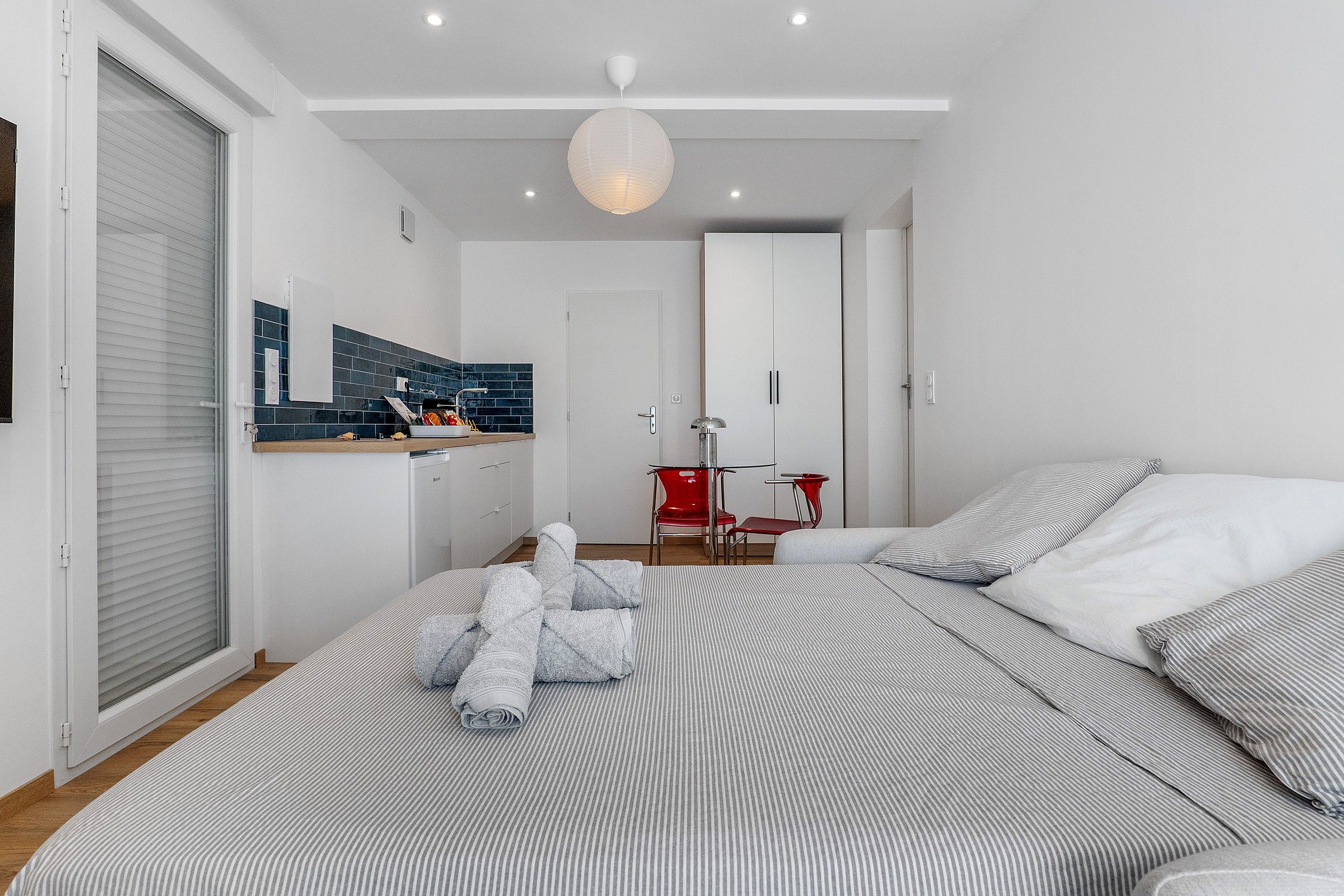 Quarto moderno com cama grande, uma cadeira vermelha, guarda-roupa branco e área de cozinha com azulejos azuis.