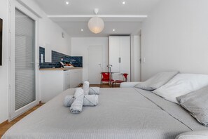 Et moderne soveværelse med en stor seng, en rød stol, et hvidt skab og et køkkenområde med blå fliser.
