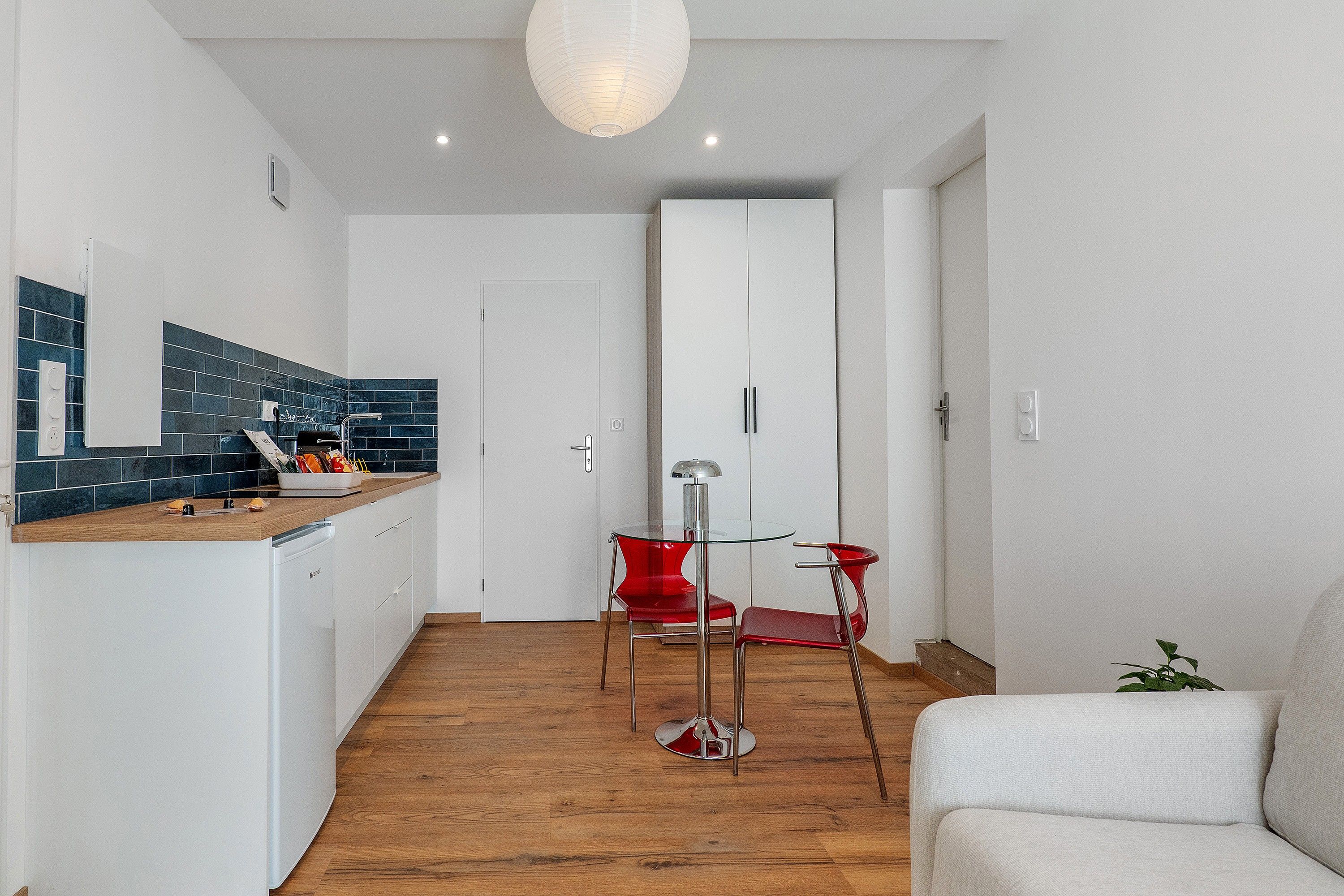 Une cuisine moderne avec un plan de travail en bois, un coin repas avec une table en verre et des chaises rouges, et une armoire blanche en arrière-plan.